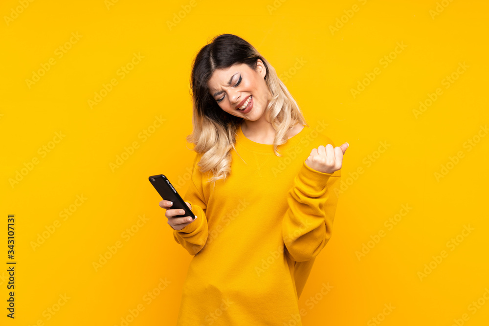 Teenager girl isolated on yellow background with phone in victory position Teenager girl isolated on yellow background with phone in victory position