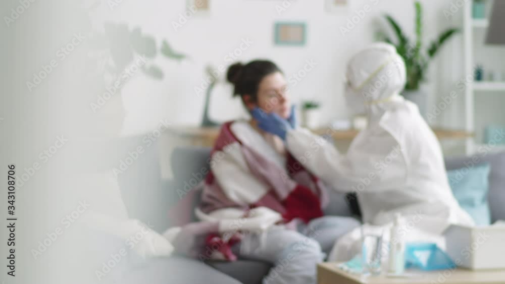 Defocused shot of doctor in protective suit sitting on couch in living room and examining female patient while giving coronavirus at-home test during isolation Defocused shot of doctor in protective suit sitting on couch in living room and examining female patient while giving coronavirus at-home test during isolation
