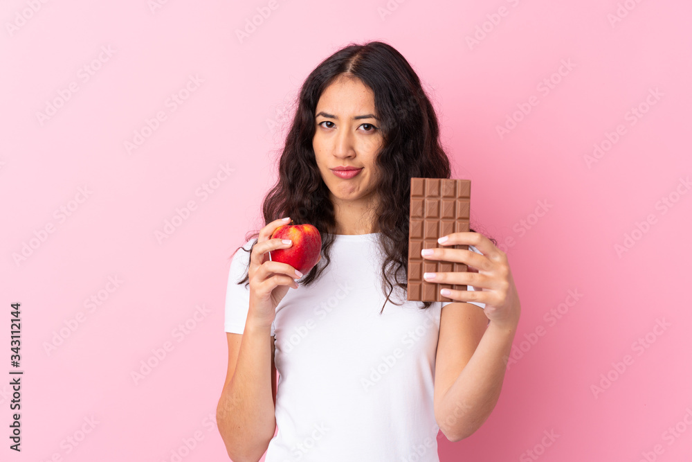 Spanish Chinese woman over isolated pink background taking a chocolate tablet in one hand and an apple in the other Spanish Chinese woman over isolated pink background taking a chocolate tablet in one hand and an apple in the other
