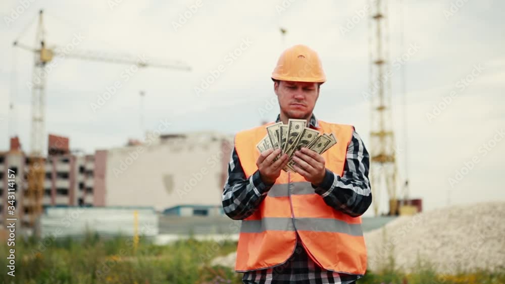 A man in an orange vest and orange helmet looks at the money and then throws it in different directions against the background of a construction site. A man in an orange vest and orange helmet looks at the money and then throws it in different directions against the background of a construction site.
