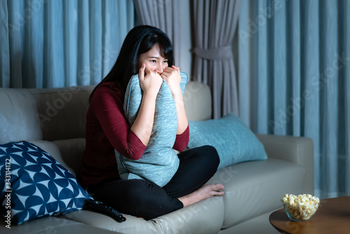 Фотография Young asian woman watching television suspense movie or news looking shocked and excited eating popcorn late night at home living room couch