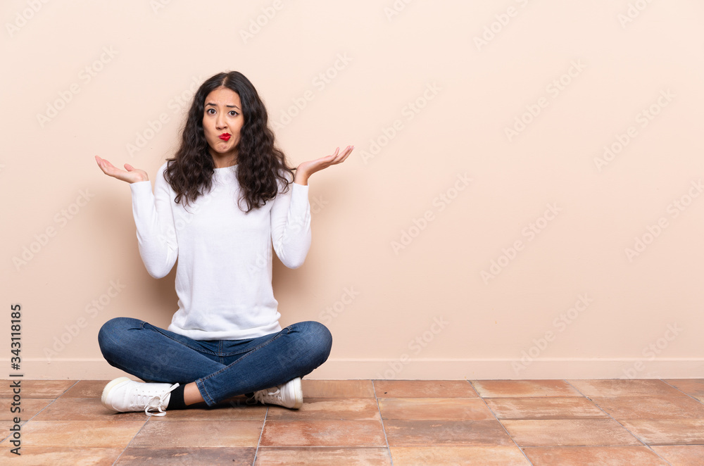 Young woman sitting on the floor having doubts while raising hands Young woman sitting on the floor having doubts while raising hands