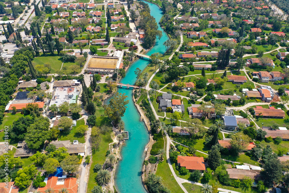 Aerial image of Kibbutz Nir David with Amal river channel turquoise water dividing east and west side riverside houses and palm trees, Israel.