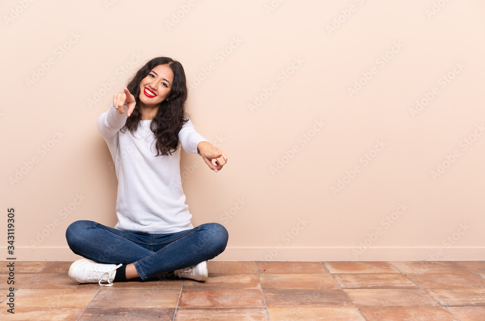 Young woman sitting on the floor points finger at you while smiling Young woman sitting on the floor points finger at you while smiling