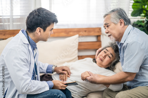 Asian senior old woman with bad condition feeling sick lying on sofa having medical check up by doctor visiting at home. Elder man husband taking care of wife closely and worry about unwell illness.