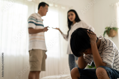 Asian boy kid sitting and crying on bed while parents having fighting or quarrel conflict at home. Child covering face and eyes with hands do not want to see the violence. Domestic problem in family.