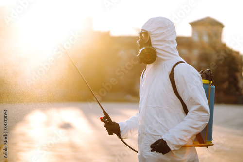 A man wearing special protective disinfection suit sprays sterilizer  in the empty public place at dawn in the city of quarantine. Covid -19. Cleaning concept. © maxbelchenko