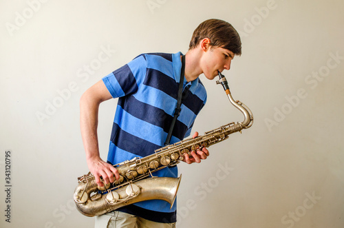 Fotografie Young saxophonist plays tenor saxophone in a striped blue shirt on a white backg