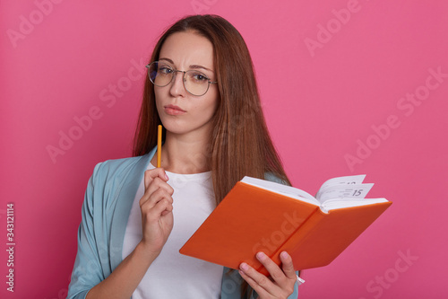 Image of thoughtful smart female wearing spectacles, holding orange notebook and pen, planning her time, using organizer, standing isolated over pink background in studio. Time management concept.