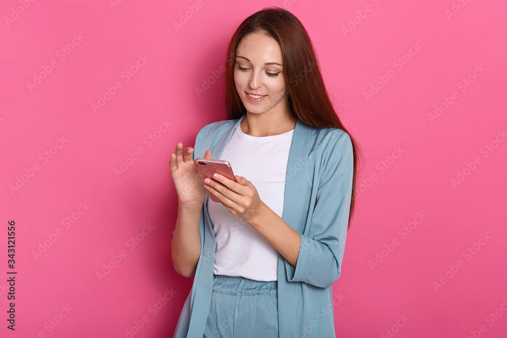 Horizontal shot of charming cheerful young lady smiling sincerely, using her device, holding smartphone, checking social networking sites, wearing smart clothes. People and social networks concept. Horizontal shot of charming cheerful young lady smiling sincerely, using her device, holding smartphone, checking social networking sites, wearing smart clothes. People and social networks concept.