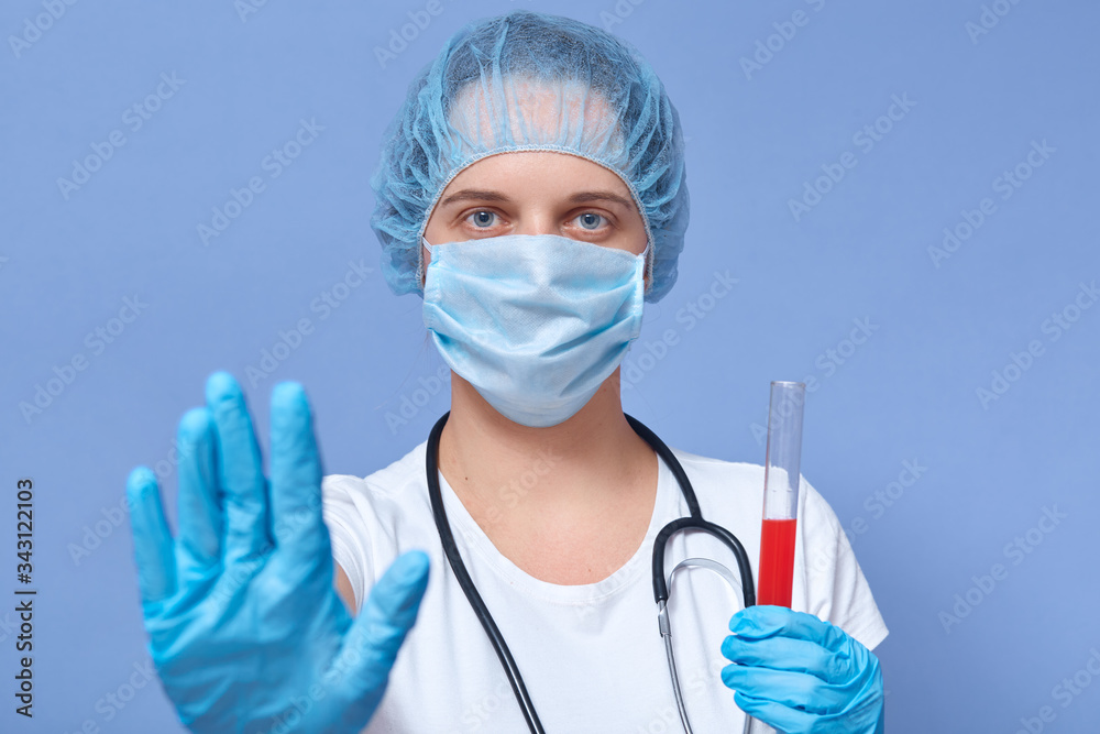 Picture of serious hard working doctor holding tube with blood for coronavirus test, making gesture, raising hand, wearing protective mask, hat and gloves, controling situation. Covid19 concept. Picture of serious hard working doctor holding tube with blood for coronavirus test, making gesture, raising hand, wearing protective mask, hat and gloves, controling situation. Covid19 concept.