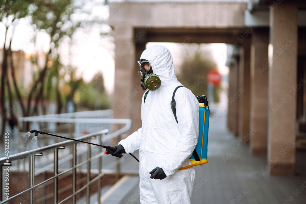 Man in protective suit and mask sprays disinfector onto the railing in the empty public place at dawn in the city of quarantine. Covid -19. Cleaning concept. Man in protective suit and mask sprays disinfector onto the railing in the empty public place at dawn in the city of quarantine. Covid -19. Cleaning concept.