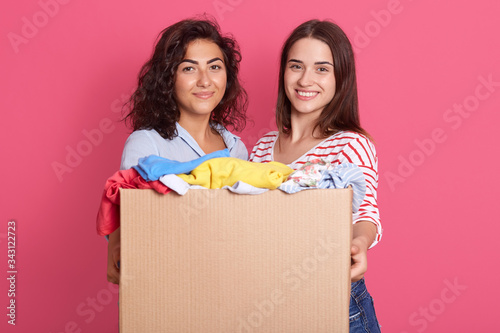 Closeup portrait of two adorable females with dark hair, looking at camera with smiles, holding box full of clothing for poor and homeless people, ladies like helping. Charity and donation concept. photo