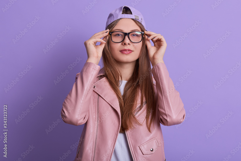 Studio shot of young lady wearing rosy jacket,hat and eyewears, woman keeping hands on frame of glasses, looking at camera, standing with calm facial expression against lilac wall.People concept. Studio shot of young lady wearing rosy jacket,hat and eyewears, woman keeping hands on frame of glasses, looking at camera, standing with calm facial expression against lilac wall.People concept.