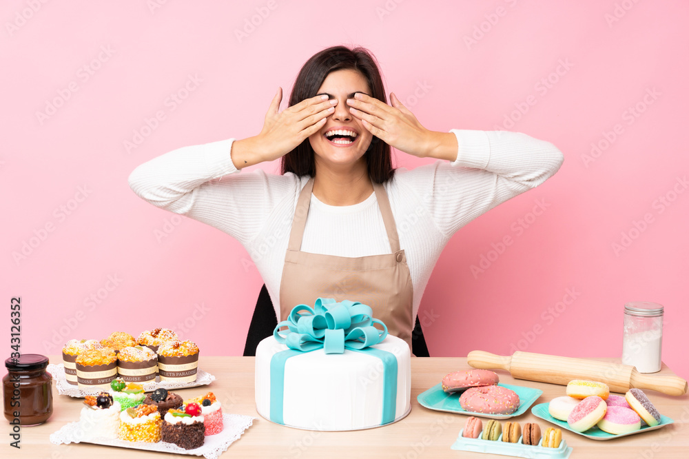 Pastry chef with a big cake in a table over isolated pink background covering eyes by hands Pastry chef with a big cake in a table over isolated pink background covering eyes by hands