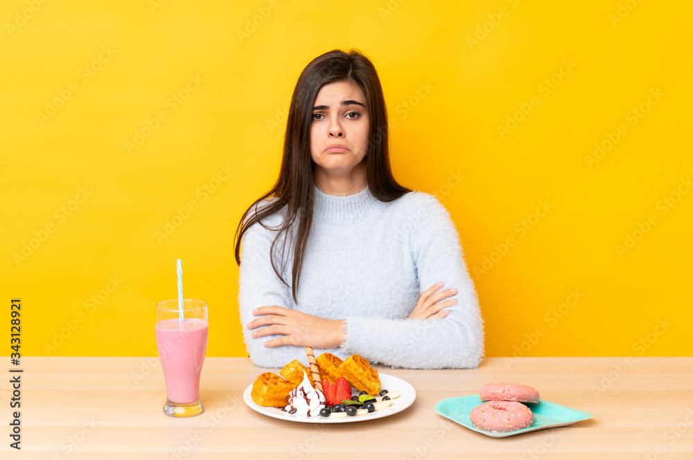 Young woman eating waffles and milkshake in a table over isolated yellow background sad Young woman eating waffles and milkshake in a table over isolated yellow background sad