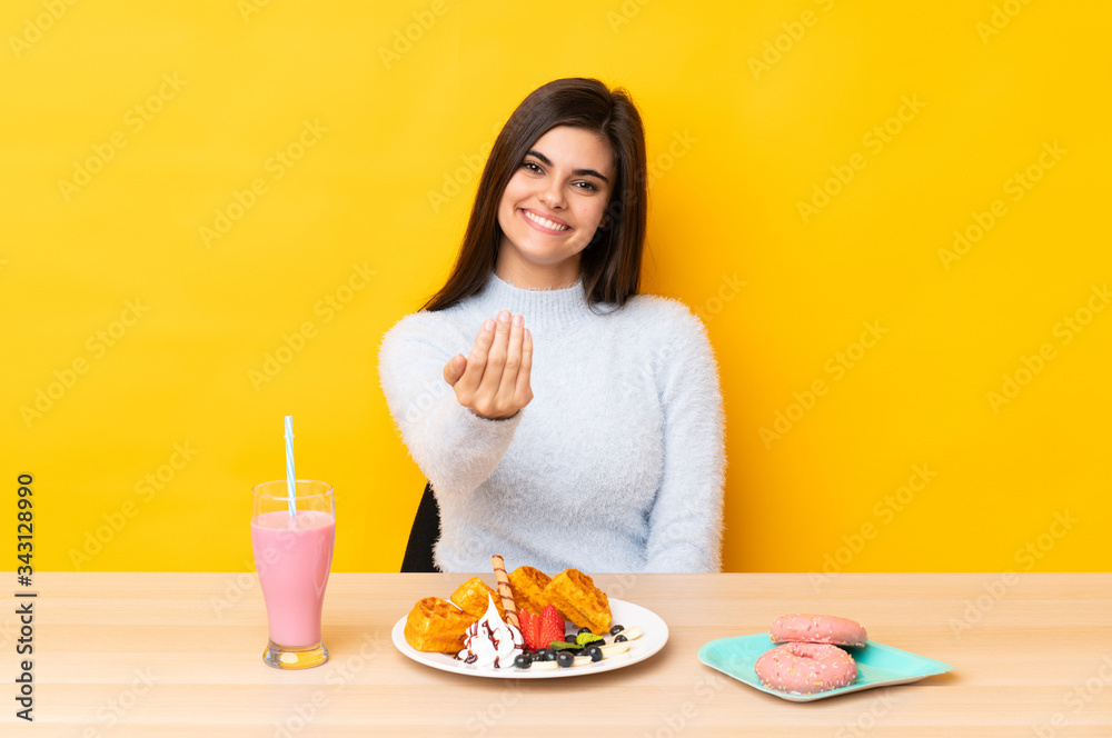 Young woman eating waffles and milkshake in a table over isolated yellow background inviting to come Young woman eating waffles and milkshake in a table over isolated yellow background inviting to come