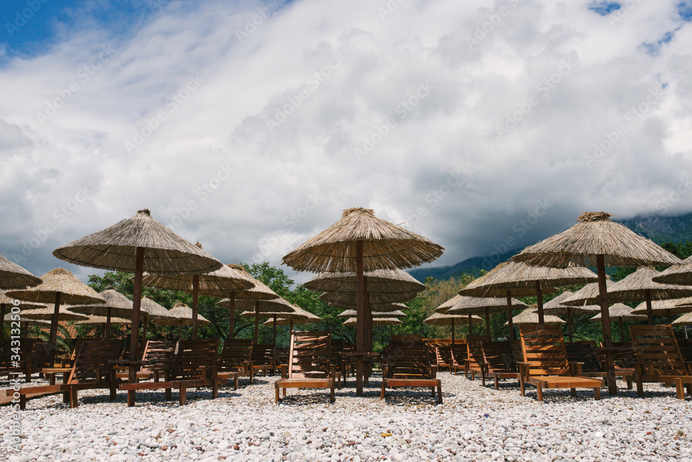 Sunbeds and umbrellas on a deserted pebble beach in cloudy weather on a background of clouds in Budva, Montenegro.