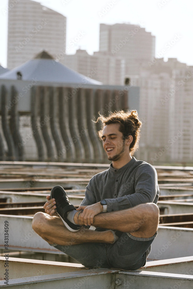 Positive young european man crossing legs in lotus pose croosed legs on blurry urban residential building city background. Urban yoga concept. Relaxation in city life Positive young european man crossing legs in lotus pose croosed legs on blurry urban residential building city background. Urban yoga concept. Relaxation in city life