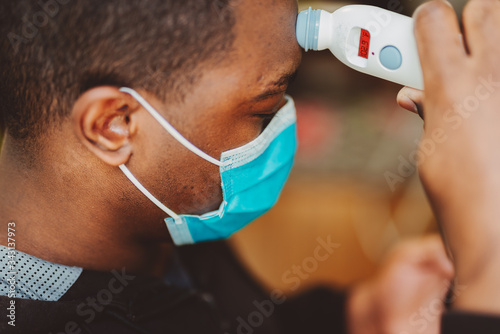 African american man using a temporal thermometer to record his temperature by running it across his forehead, man wearing surgical mask