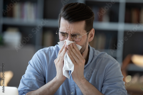 Close up unhealthy businessman blowing wiping running nose, using napkin, sick young man wearing glasses feeling unwell, coughing, seasonal allergy or respiratory disease symptoms concept
