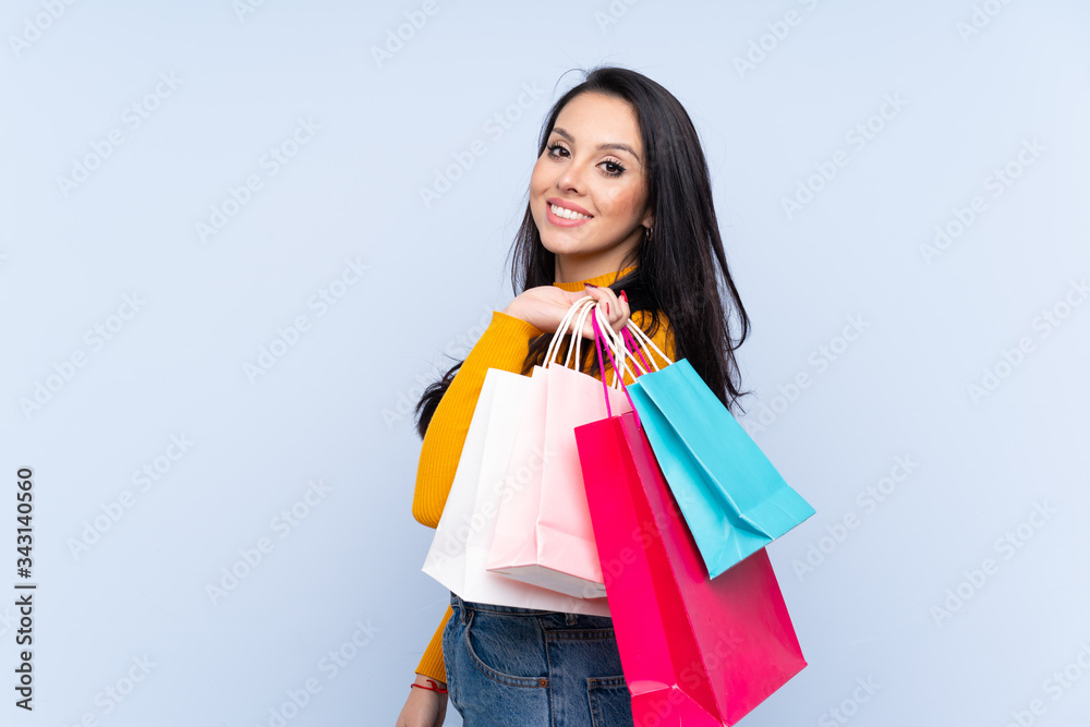 Young Colombian girl over isolated blue background holding shopping bags and smiling