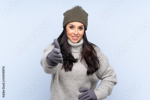 Young Colombian girl with winter hat over isolated blue background handshaking after good deal