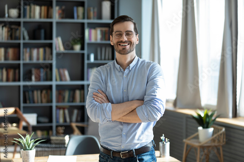 Portrait laughing businessman with arms crossed standing near work desk in modern cabinet, looking at camera, happy excited entrepreneur wearing glasses posing for photo at workplace