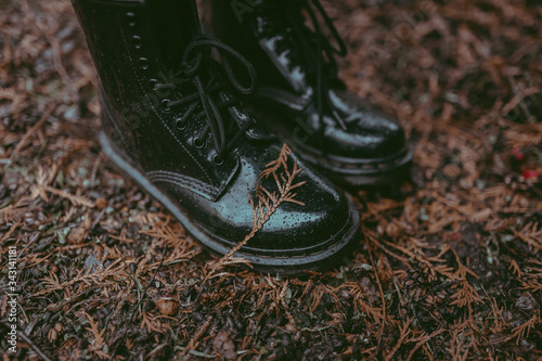A close-up of black rubber waterproof boots with laces and rain drops and fallen dry leaf on the smooth shiny material. A ground covered with orange leaves. An autumn walk in a park in rain boots