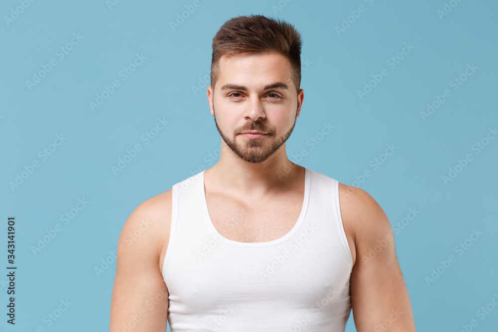 Handsome smiling young bearded guy 20s in white singlet posing isolated on pastel blue wall background studio portrait. Sport fitness healthy lifestyle concept. Mock up copy space. Looking camera. Handsome smiling young bearded guy 20s in white singlet posing isolated on pastel blue wall background studio portrait. Sport fitness healthy lifestyle concept. Mock up copy space. Looking camera.