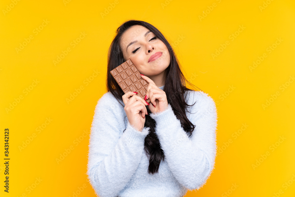 Young Colombian girl over isolated yellow background taking a chocolate tablet and happy Young Colombian girl over isolated yellow background taking a chocolate tablet and happy