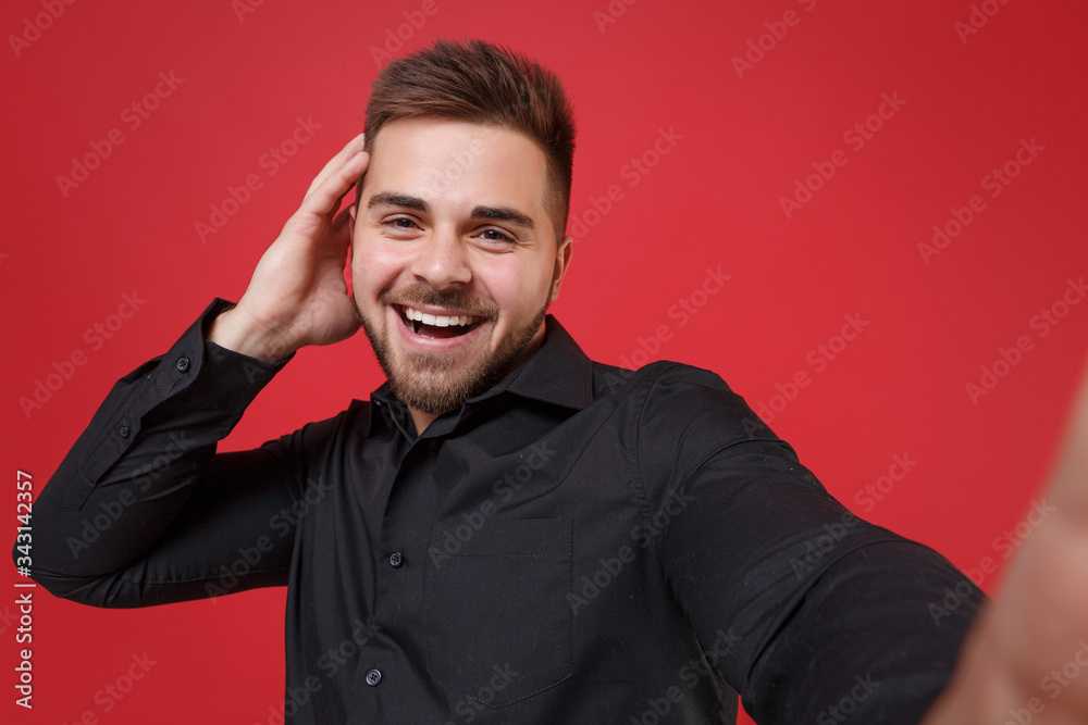 Close up of cheerful young bearded guy 20s in classic black shirt posing isolated on red background. People lifestyle concept. Mock up copy space. Doing selfie shot on mobile phone, put hand on head. Close up of cheerful young bearded guy 20s in classic black shirt posing isolated on red background. People lifestyle concept. Mock up copy space. Doing selfie shot on mobile phone, put hand on head.