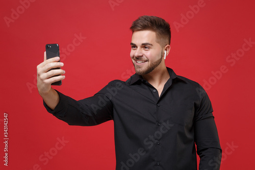 Smiling young bearded guy in classic black shirt posing isolated on red background. People lifestyle concept. Mock up copy space. Using air pods doing selfie shot or making video call on mobile phone.