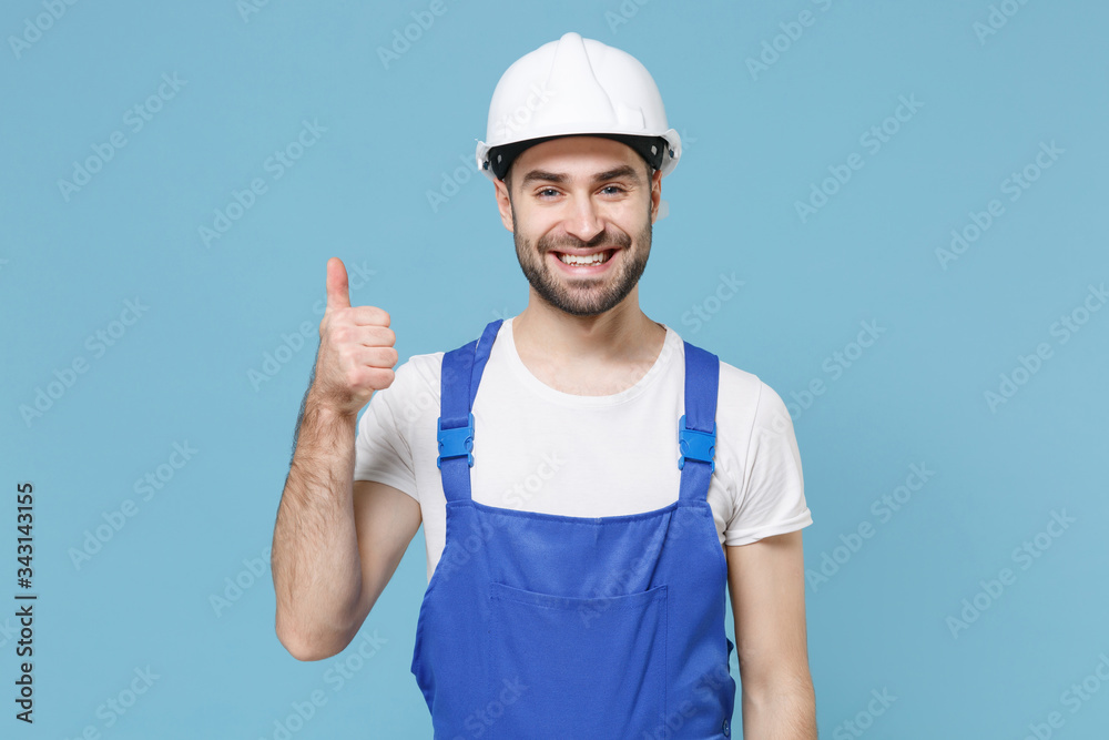 Smiling young man in coveralls protective helmet hardhat isolated on blue wall background studio portrait. Instruments accessories for renovation apartment room. Repair home concept. Showing thumb up.