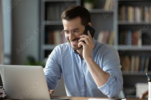 Smiling businessman talking on cellphone, making call, looking at laptop screen, friendly manager consulting customer by phone, happy man chatting with friends, having pleasant conversation photo