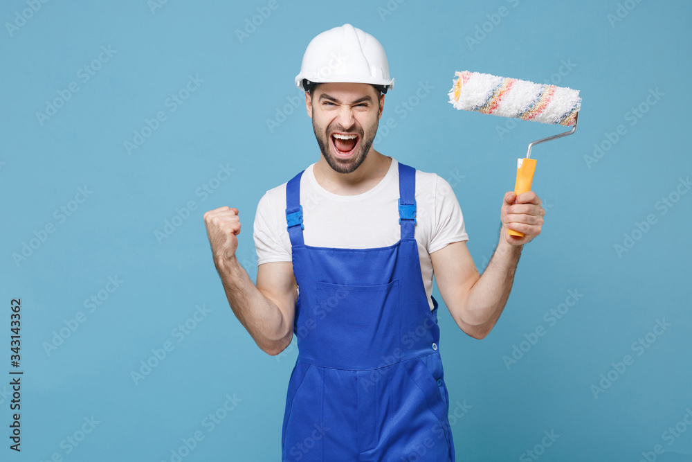 Overjoyed man in coveralls protective helmet hardhat hold paint roller isolated on blue background. Instruments accessories for renovation apartment room. Repair home concept. Doing winner gesture.
