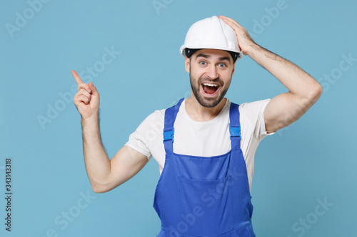 Excited young man in coveralls helmet hardhat isolated on blue background. Instruments accessories for renovation apartment room. Repair home concept. Pointing index finger aside up, put hand on head.