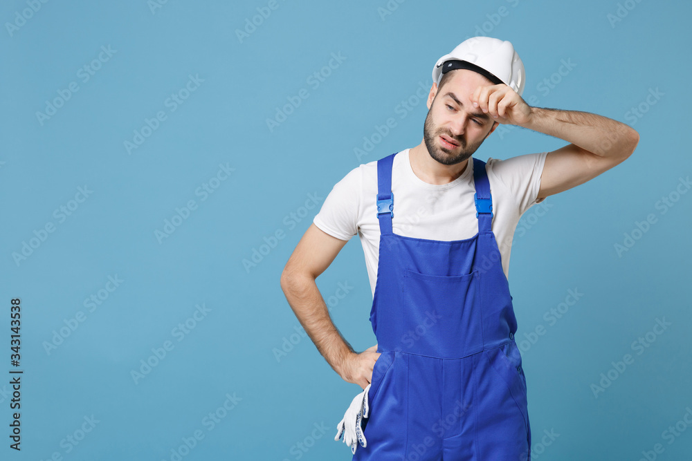 Puzzled tired young man in coveralls protective helmet hardhat isolated on blue wall background studio. Instruments accessories for renovation apartment room. Repair home concept. Put hand on head.