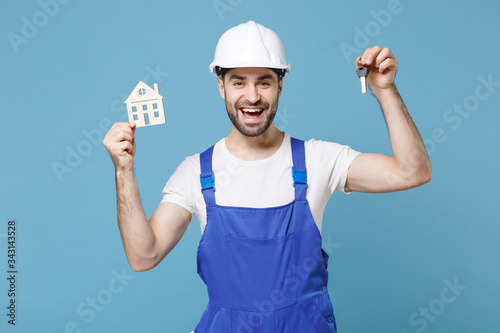 Cheerful young man in coveralls protective helmet hardhat hold house bunch of keys isolated on blue background in studio. Instruments accessories for renovation apartment room. Repair home concept.