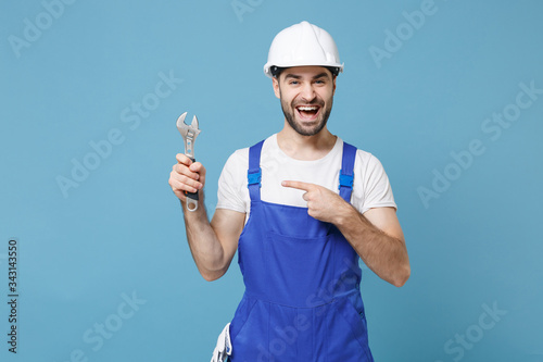 Laughing man in coveralls protective helmet hardhat pointing index finger on adjustable wrench isolated on blue background. Instruments accessories for renovation apartment room. Repair home concept.