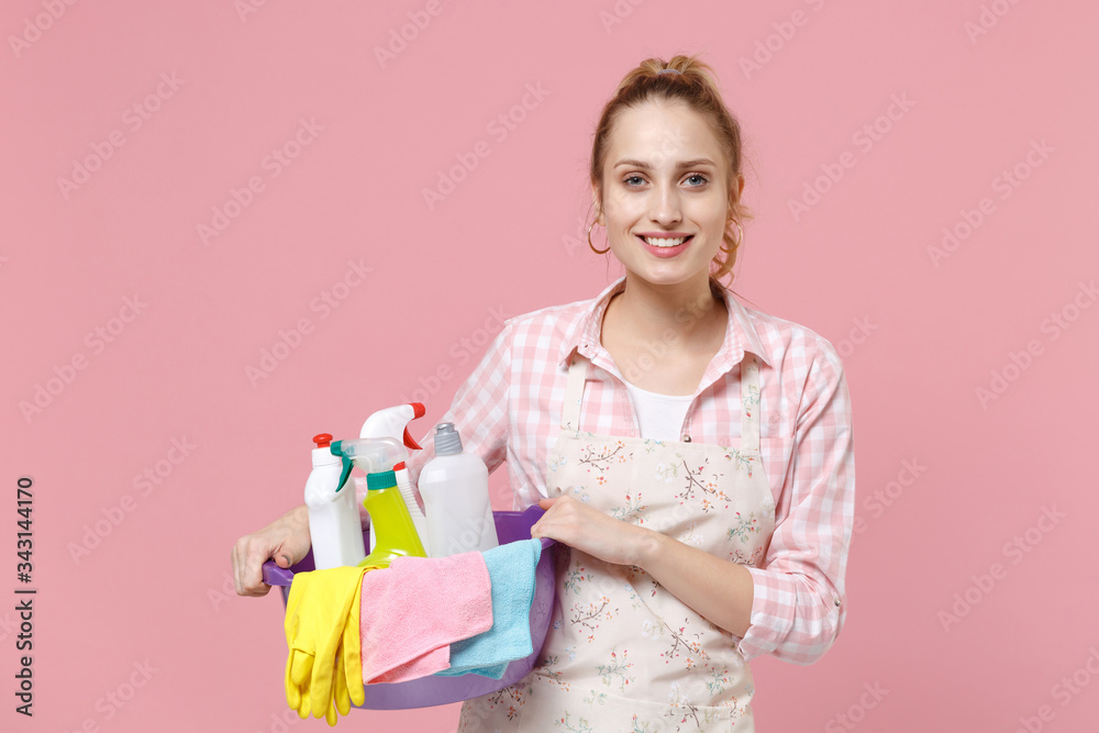 Smiling young woman housewife in apron hold basin with detergent bottles washing cleansers while doing housework isolated on pink background studio portrait. Housekeeping concept. Looking camera. Smiling young woman housewife in apron hold basin with detergent bottles washing cleansers while doing housework isolated on pink background studio portrait. Housekeeping concept. Looking camera.