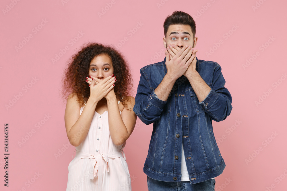 Shocked young couple friends european guy african american girl in casual clothes isolated on pastel pink background in studio. People lifestyle concept. Mock up copy space. Covering mouth with hands. Shocked young couple friends european guy african american girl in casual clothes isolated on pastel pink background in studio. People lifestyle concept. Mock up copy space. Covering mouth with hands.