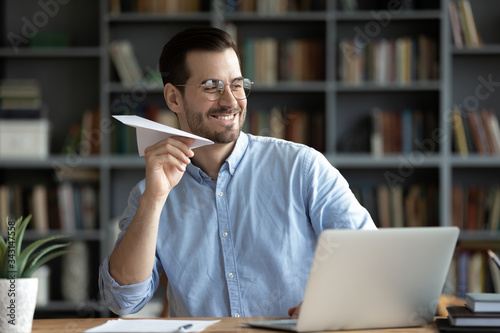 Happy smiling businessman wearing glasses playing with paper plane at workplace, sitting at desk, having fun during break, enjoying rest after work done, new opportunities and success