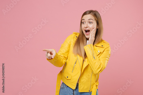 Amazed young woman girl in yellow leather jacket posing isolated on pastel pink background studio portrait. People lifestyle concept. Mock up copy space. Pointing index finger aside put hand on cheek.