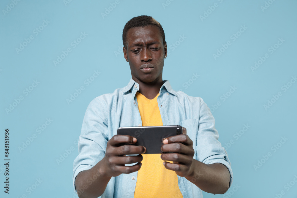 Displeased young african american man guy in casual shirt, yellow t-shirt posing isolated on blue wall background in studio. People lifestyle concept. Mock up copy space. Play game with mobile phone. Displeased young african american man guy in casual shirt, yellow t-shirt posing isolated on blue wall background in studio. People lifestyle concept. Mock up copy space. Play game with mobile phone.