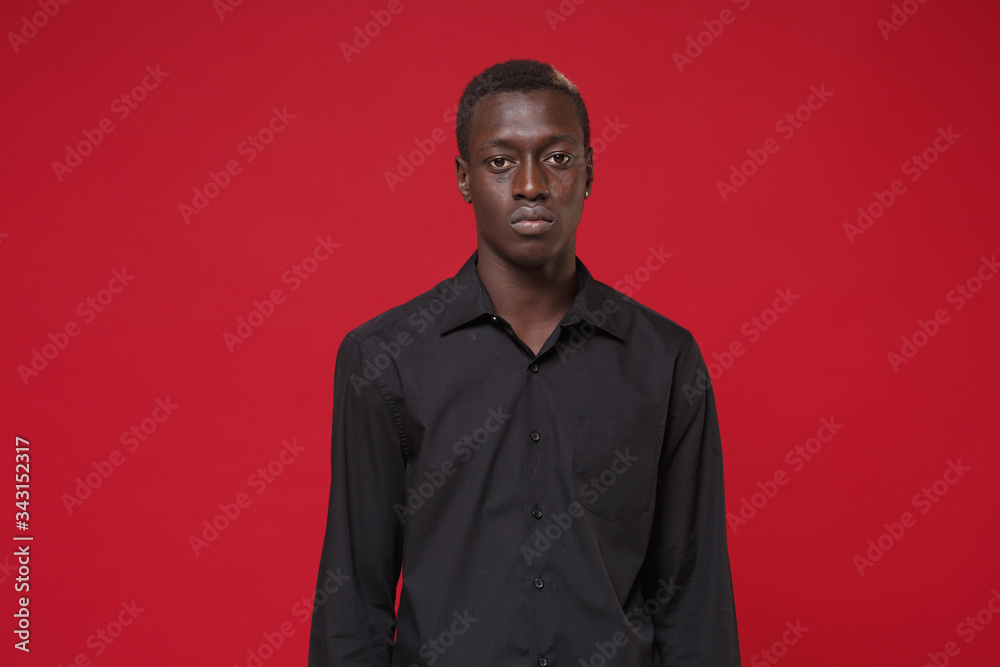 Serious young african american man guy in classic black shirt posing isolated on red wall background studio portrait. People sincere emotions lifestyle concept. Mock up copy space. Looking camera. Serious young african american man guy in classic black shirt posing isolated on red wall background studio portrait. People sincere emotions lifestyle concept. Mock up copy space. Looking camera.