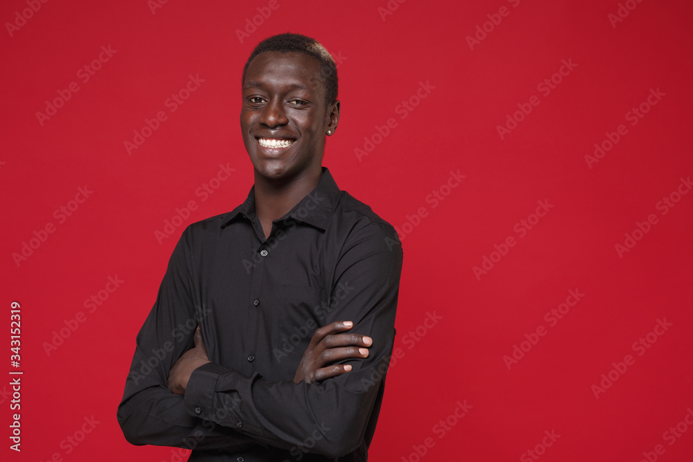 Smiling young african american man guy in classic black shirt posing isolated on red background studio portrait. People sincere emotions lifestyle concept. Mock up copy space. Holding hands crossed.