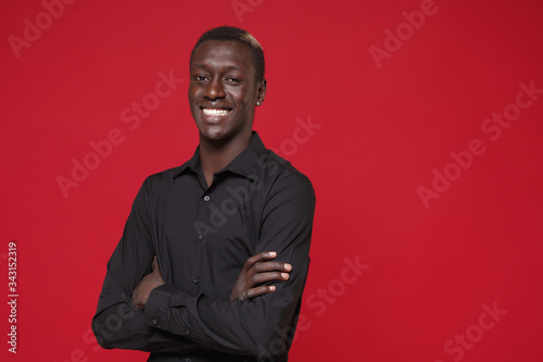 Smiling young african american man guy in classic black shirt posing isolated on red background studio portrait. People sincere emotions lifestyle concept. Mock up copy space. Holding hands crossed.