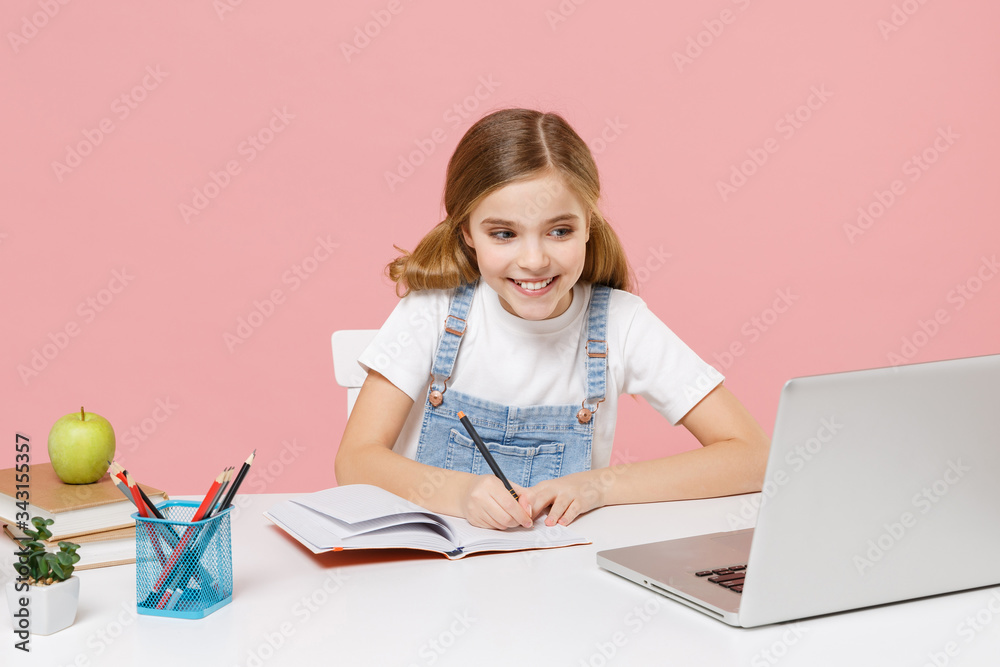 Smiling little kid schoolgirl 12-13 years old sit studying at white desk with pc laptop isolated on pink background. School distance education at home during quarantine concept. Writing in notebook. Smiling little kid schoolgirl 12-13 years old sit studying at white desk with pc laptop isolated on pink background. School distance education at home during quarantine concept. Writing in notebook.