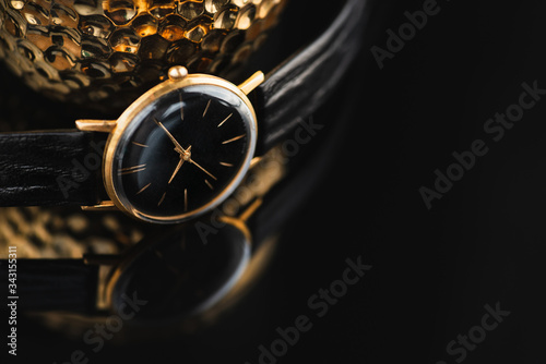 Golden wristwatch with leather strap laying on black glossy surface next to golden jewell box . Copy space.